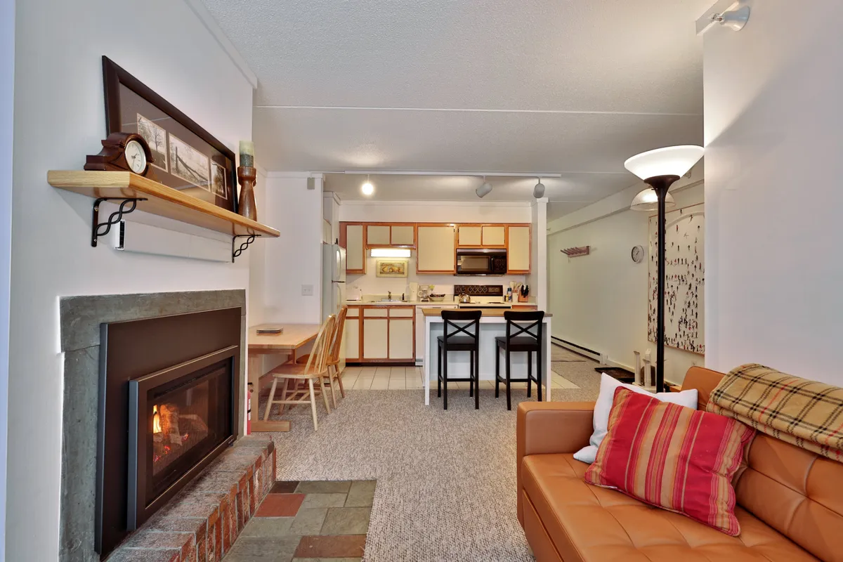 The photo centers on the living room, with a prominent fireplace (though not operational), a plush sofa, and the kitchen island in full view. The inviting mood is set by the warm tones and cozy furnishings, emphasizing areas where guests can relax, cook, and socialize.
