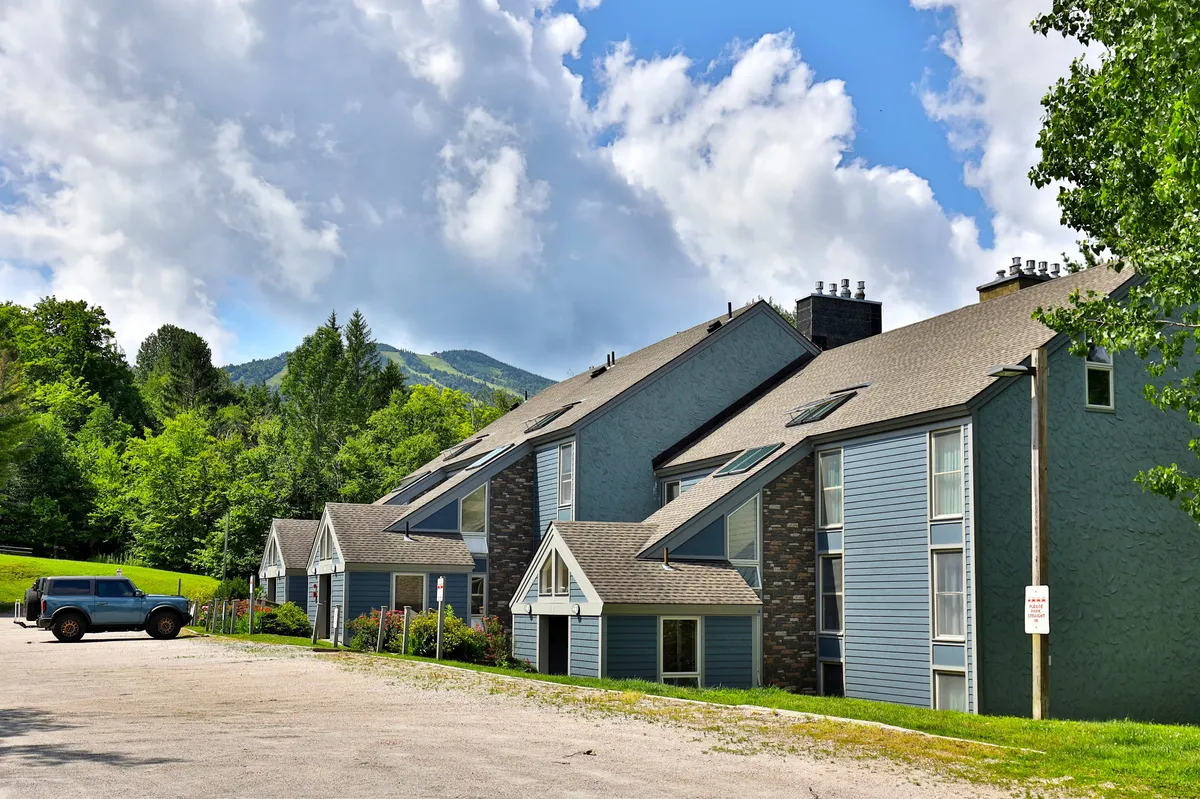 This image features the Whiffletree multi-story building nestled in a lush green landscape, with mountains rising in the background and a vehicle parked nearby. The scene captures a sense of serenity and connection to nature, with ample space for outdoor exploration and family activities.