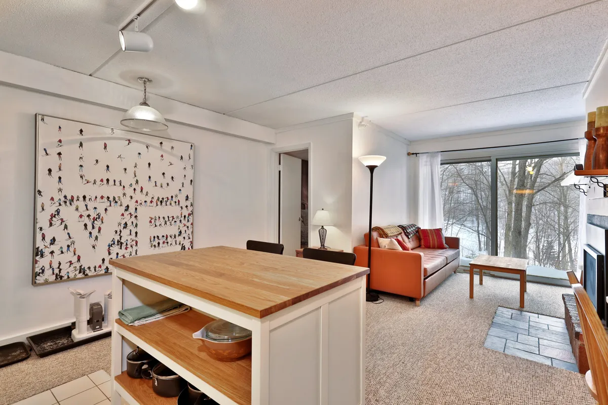 This image showcases the main living area featuring a comfortable orange sofa, a large piece of artwork on the wall, a wooden dining table, and part of the kitchen. Sunlight streams in from large windows, creating a warm, inviting atmosphere. The open layout connects the living, dining, and kitchen spaces, fostering a sense of togetherness.