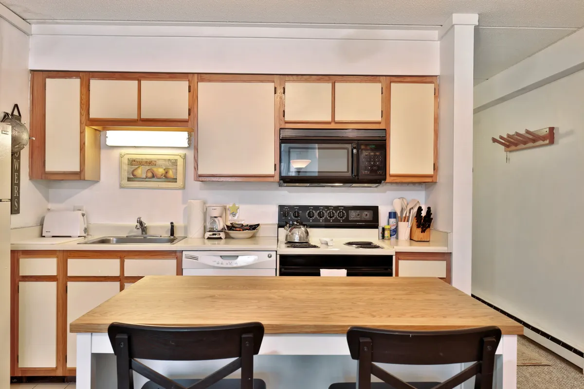 A warm, welcoming kitchen and dining area featuring a wooden table with seating, visible appliances, and colorful artwork on the walls. The space is well-lit and designed for group meals and casual gatherings.