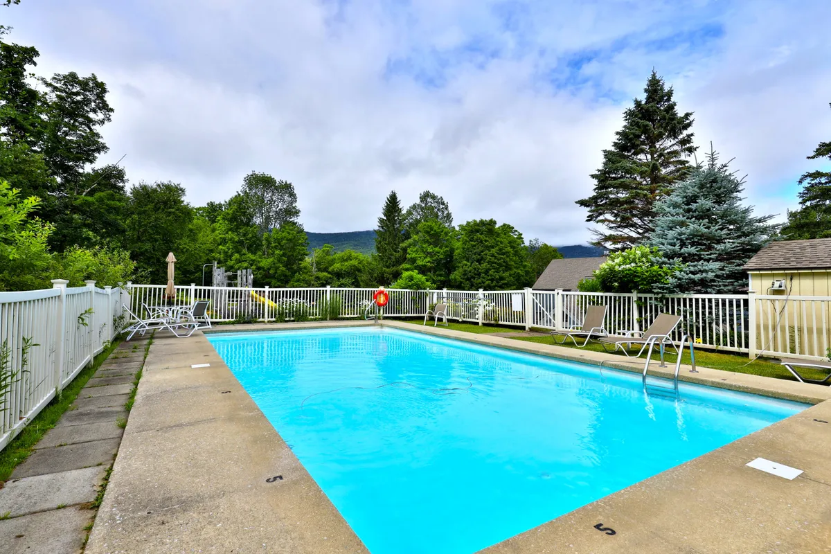 A sparkling outdoor swimming pool surrounded by lush green trees and several poolside lounge chairs. The scene conveys a relaxed, vacation-like atmosphere, perfect for summer leisure and socializing.
