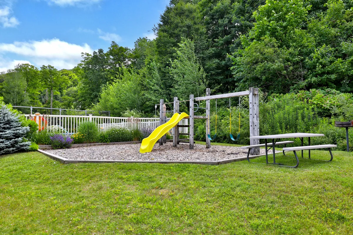 A vibrant outdoor space with a yellow slide, swing set, picnic table, and surrounding greenery. Children can be seen playing on the equipment, while families gather at the picnic table, creating an atmosphere of joyful energy and togetherness.