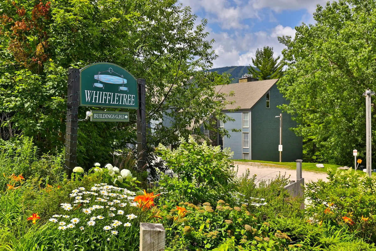 An inviting exterior shot featuring the Whiffletree sign surrounded by colorful flower beds and abundant greenery, with the building structure in the background. The lush garden reflects seasonal changes and offers a welcoming first impression.