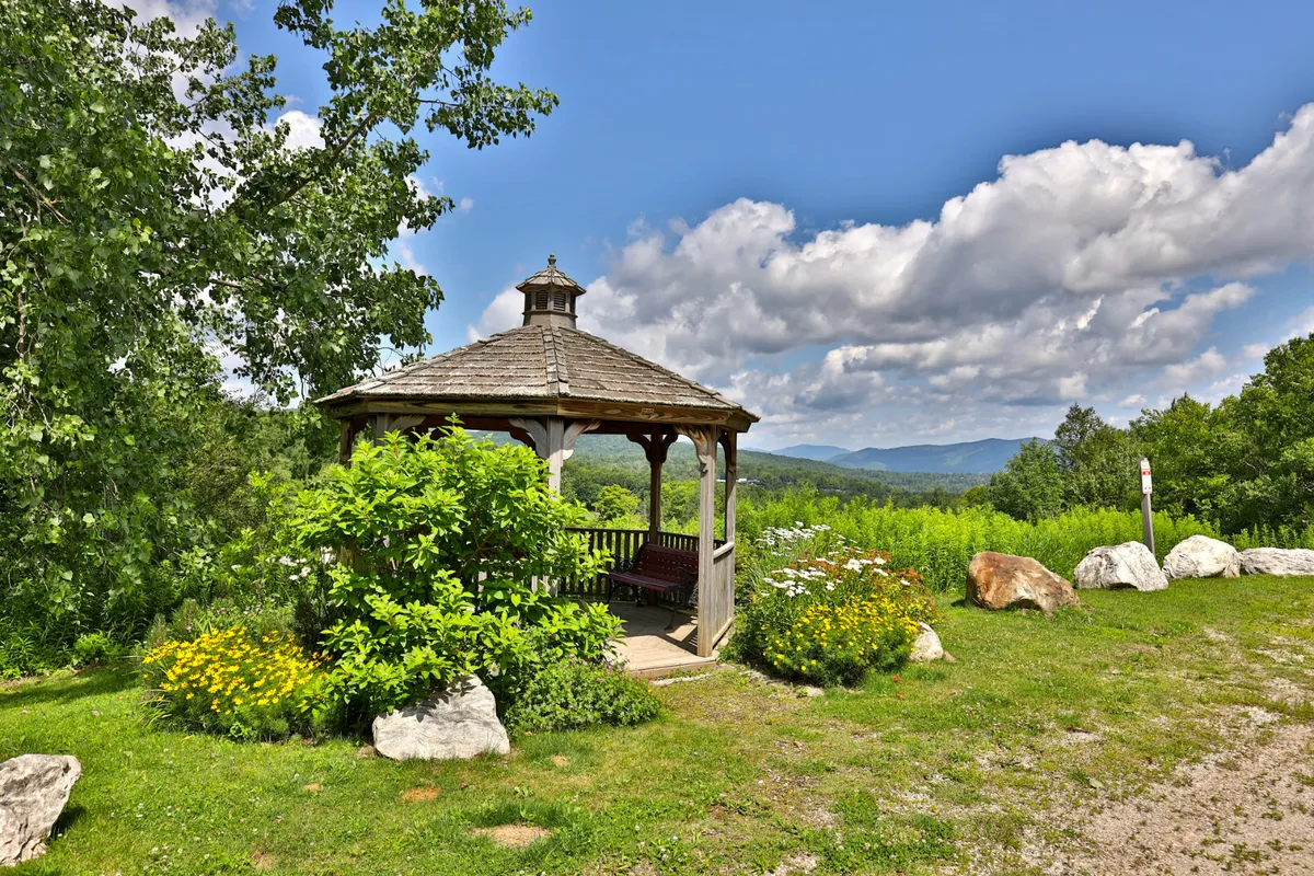 This exterior image features a charming gazebo surrounded by blooming plants, set against the backdrop of distant mountains. The tranquil scene captures a lush garden area perfect for relaxation and quiet contemplation. The inviting setting reflects both a communal and private space for enjoying the natural surroundings.