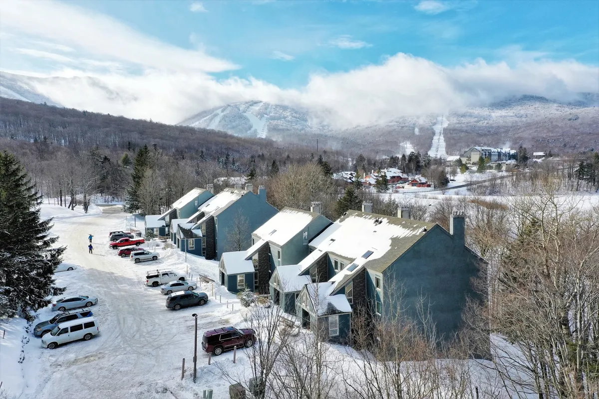 A tranquil scene of snow-covered rooftops framed by a tree line, with the mountains rising in the distance and parked vehicles suggesting guest arrivals. The ambiance is peaceful yet active, hinting at outdoor winter activities and the convenience of slope-side stays.