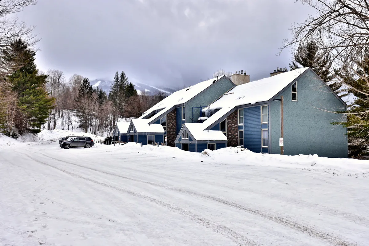 A snowy scene with charming, snow-covered buildings, surrounding trees, and a parked car, conveying the cozy atmosphere of a mountain town in winter. The image subtly suggests both adventure and the comfort of returning 'home' afterward.
