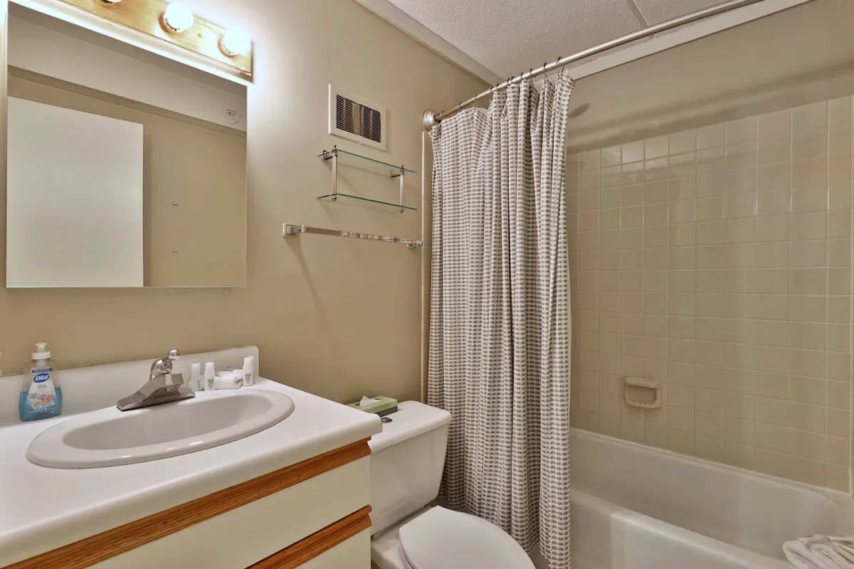 A clean, functional bathroom with a sink, mirror, shower curtain, toilet, and bath tub. The neutral colors and tidy layout suggest freshness, while the presence of linens and towels ensures guest comfort.