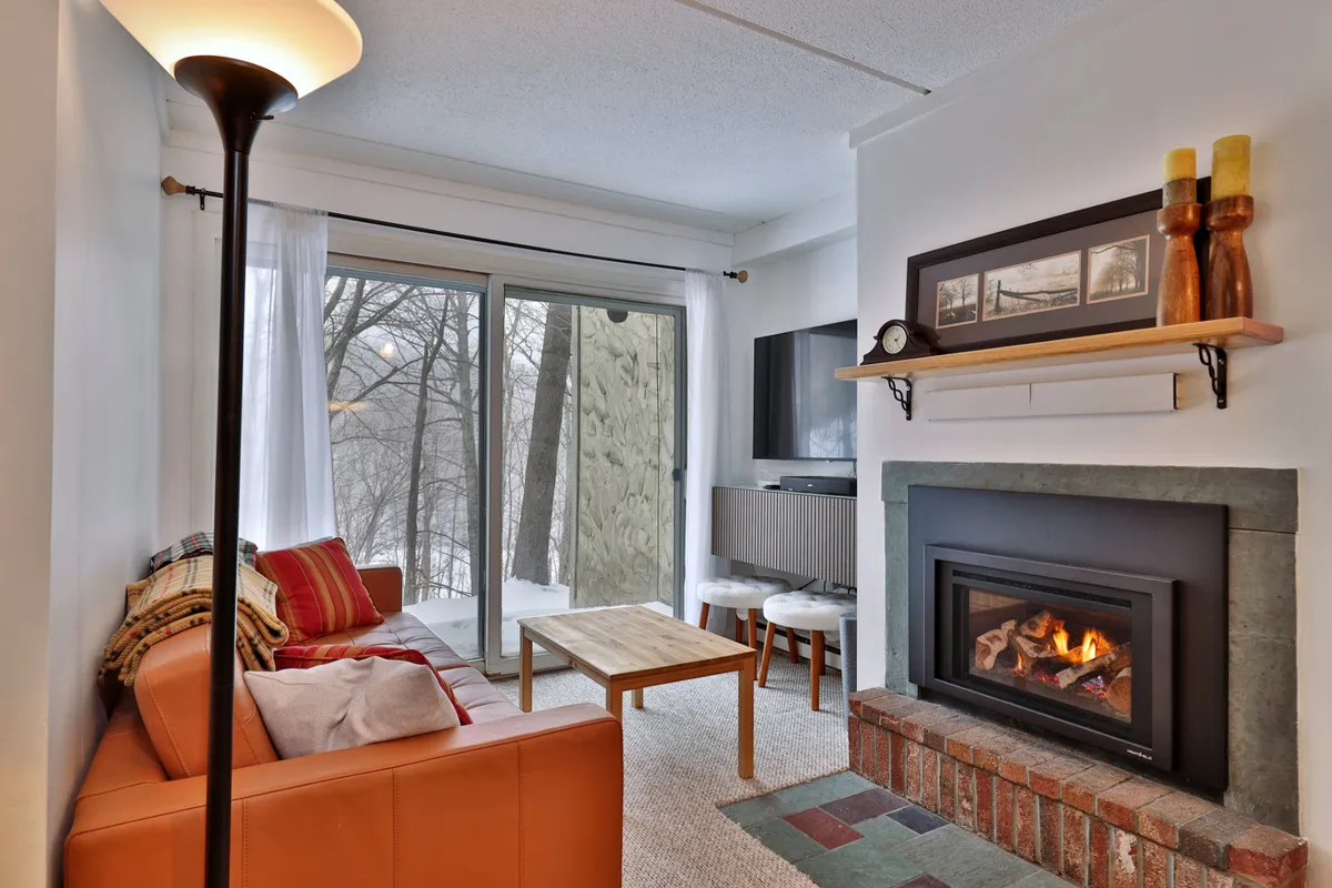 The image showcases a warm, inviting living room anchored by a glowing fireplace, a plush sofa perfect for unwinding, and expansive windows revealing snowy Vermont scenery just beyond.