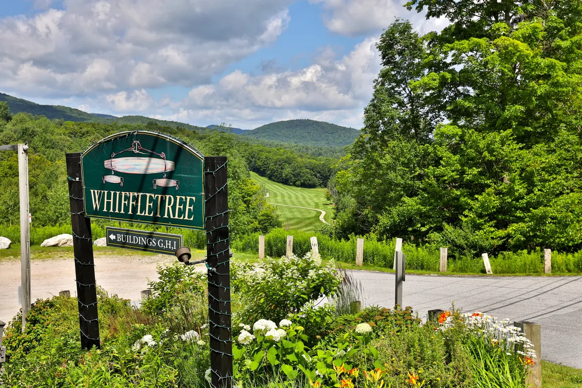 An exterior view showcasing the Whiffletree sign, lush green landscaping, and scenic mountains in the background. Well-maintained flower gardens reinforce a sense of natural beauty, while the trail-like pathways invite exploration and outdoor activity.
