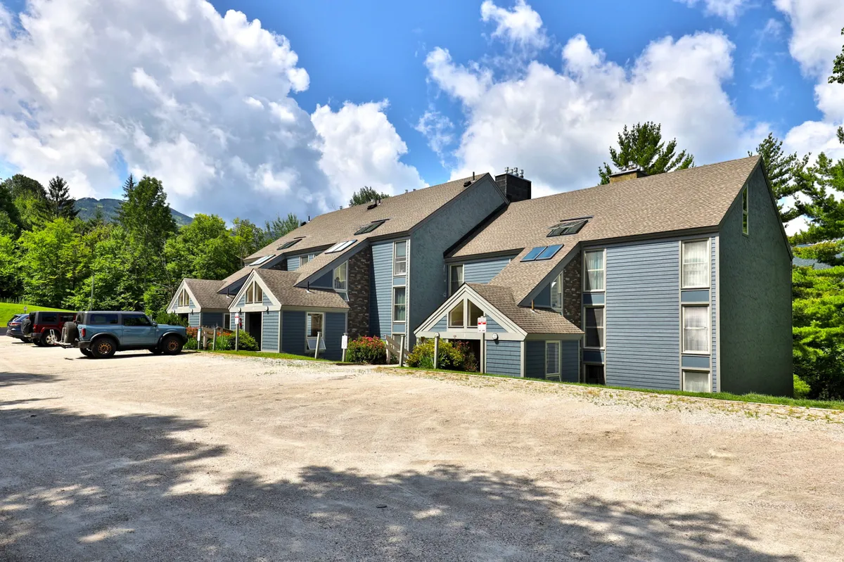 The condominium building is shown with its distinctive architecture, a generous parking area, and abundant surrounding greenery. The setting hints at a welcoming community and outdoor gathering space.