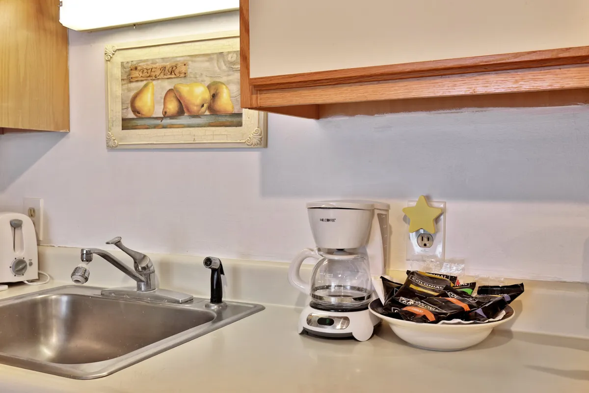A close-up of the kitchen counter featuring a coffee maker, bowl of snacks, sink, and a splash of artwork in the background. The scene is cozy and intimate, highlighting daily rituals like morning coffee or sharing treats.