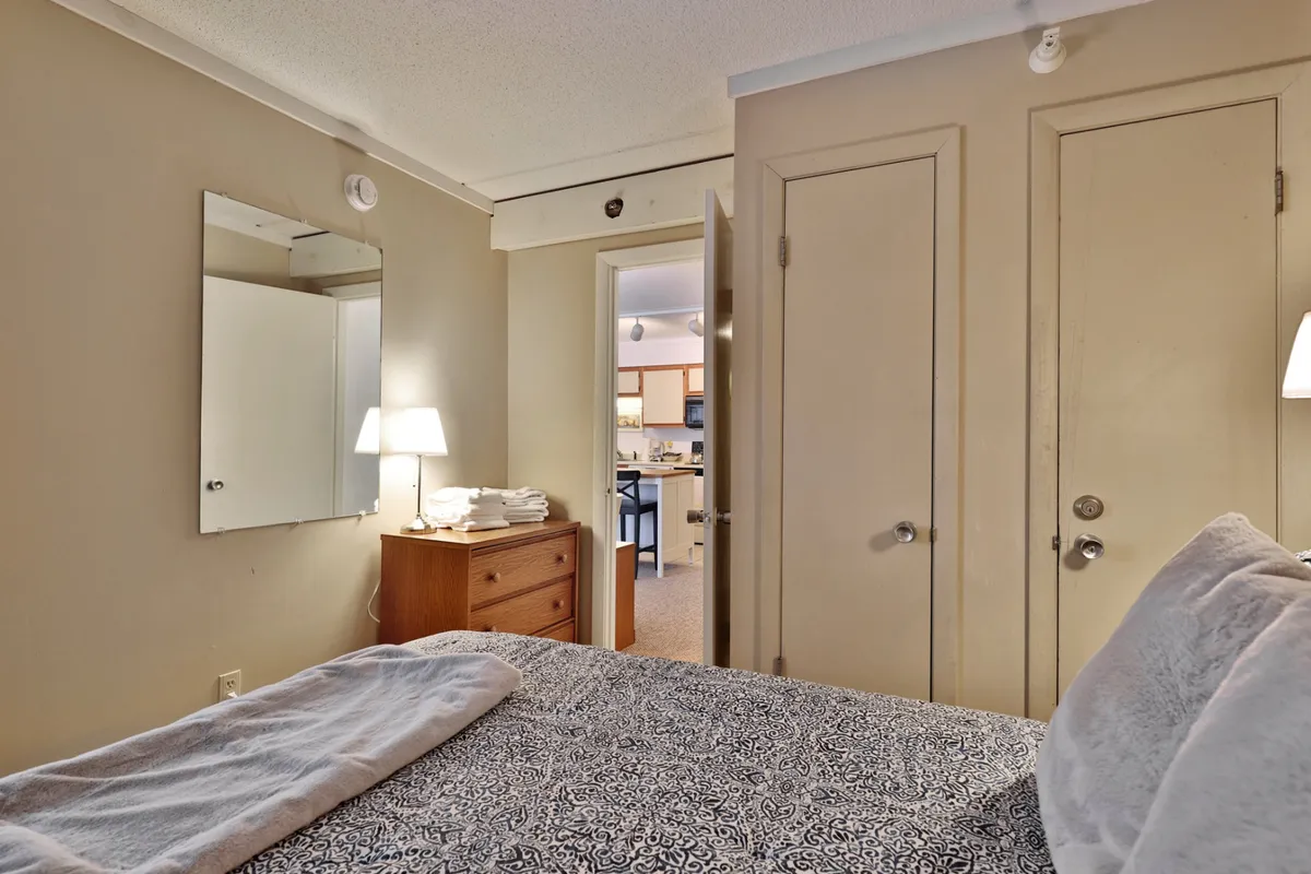 This image shows a cozy bedroom featuring a bed with patterned bedding, a nightstand with a lamp providing soft, ambient light, and a mirror that reflects brightness into the room. An open door leads to the kitchen, hinting at an accessible layout. The scene feels peaceful and ideal for restful sleep.
